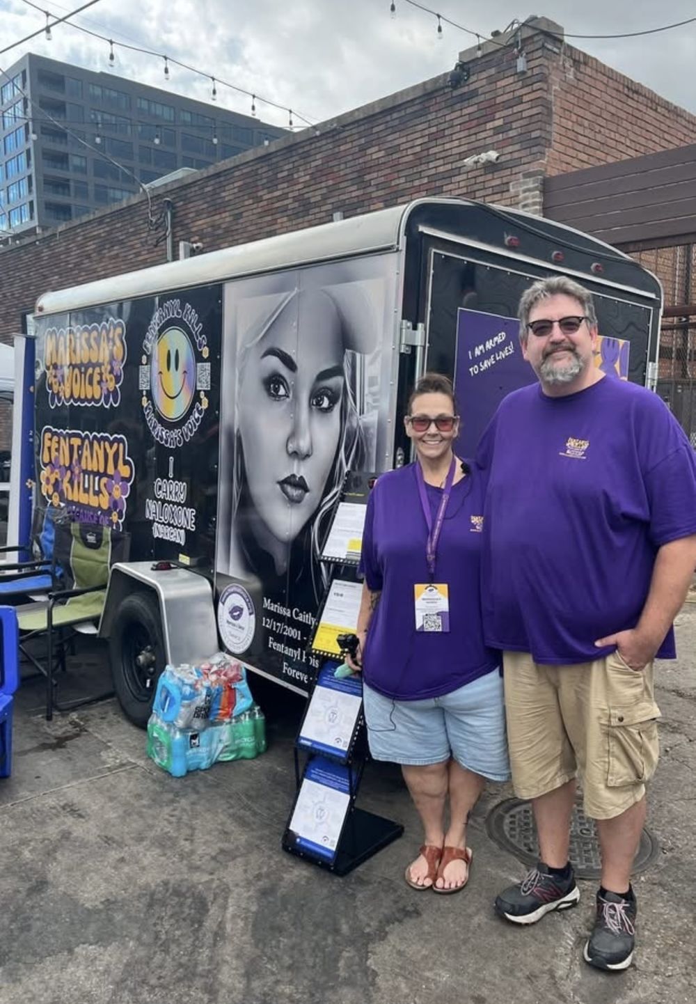 Amy and Mark standing in front of the black Marissa&rsquo;s Voice outreach trailer, which shows Marissa&rsquo;s portrait and the words Fentanyl Kills and I Carry Naloxone